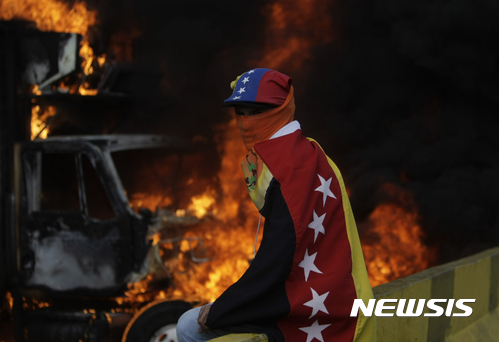 【카라카스(베네수엘라) = AP/뉴시스】 베네수엘라 국기를 몸에 두르고 복면을 한 반정부 시위대원이 27일(현지시간) 카라카스 시내에서 탈취한 트럭들을 불태우며 도로를 봉쇄하고 있다. 주말의 충돌로 또 2명의 총격 사망자가 발생, 두달째 이어져오는 시위사태의 사망자가 60명여명에 이르렀다. 2017.05.29    