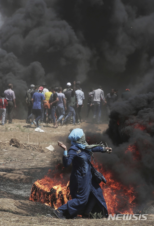【가자=AP/뉴시스】 14일 가자 지구의 이스라엘 봉쇄선 앞에서 수만 명의 팔레스타인인들이 미 대사관 예루살렘 이전에 항의하는 시위에 나선 가운데 한 여성이 이스라엘군을 향해 돌을 던지고 있다. 이 군 저격병의 시야를 가리기 위해 불 붙인 타이어에서 검은 연기가 시꺼멓게 솟아오르고 있다. 2018. 5. 14. 