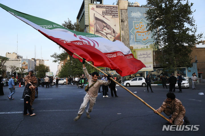 [테헤란=AP/뉴시스] 지난 15일(현지시각) 이란 테헤란의 팔레스타인 광장 건물벽에 걸린 반이스라엘 현수막 앞에서 시위대가 대형 이란 국기를 흔들고 있다. 2024.4.19