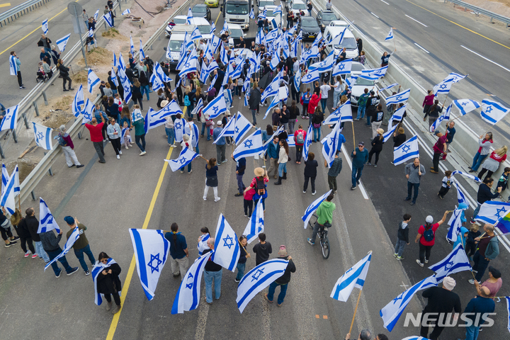 [베이트 야나이=AP/뉴시스] 23일(현지시간) 이스라엘 베이트 야나이 교차로에서 정부의 사법제도 개편에 반대하는 시위대가 국기를 흔들며 고속도로를 봉쇄하고 있다. 베냐민 네타냐후 이스라엘 총리의 사법 개혁에 반대하는 세력들이 이스라엘을 독재국가로 추락시킨다며 연일 항의 시위를 계속하는 가운데 이스라엘 의회는 23일 사법 개혁의 몇 가지 법안 중 첫 번째 법안을 통과시켰다. 2023.03.23.