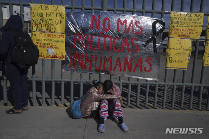  [치우다드후아레스( 멕시코)= AP/뉴시스] 멕시코 국경의 치우다드후아레스시내 이민수용소에서 화재로 40명이 숨진 뒤 베네수엘라출신의 어린 자매가 수용소 앞에서 서로 위로하며 앉아있다. "비인간적 이민정책은 이제 그만"이라고 쓴 펼침막이 보인다. 